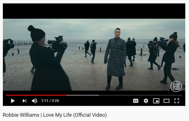 Robbie Williams in plaid coat on beach with cameras, from Love My Life video