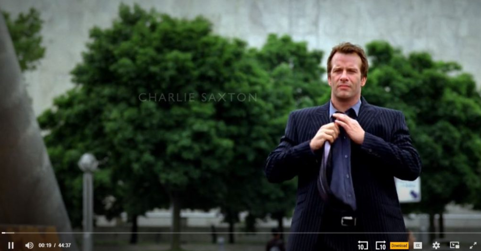 Charlie Saxton adjusts his tie in front of green trees.