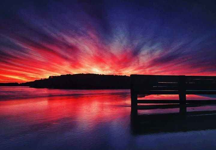 Dramatic sunset over water with silhouette of bench, reflecting holiday struggles and hope.