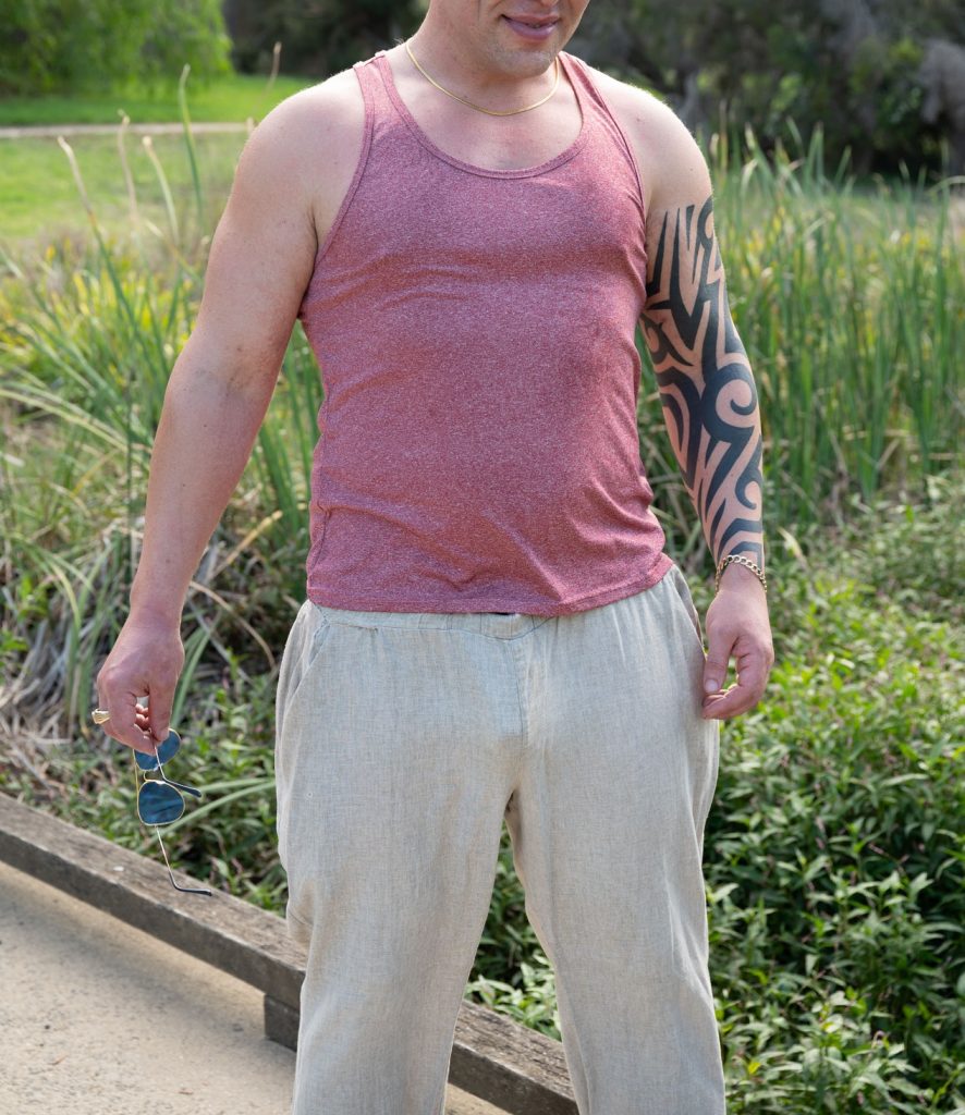 Man in tank top and linen pants holding sunglasses outdoors.