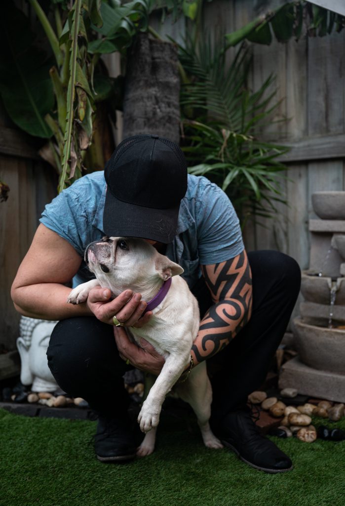 Man in black cap and blue shirt holding white French bulldog outdoors.