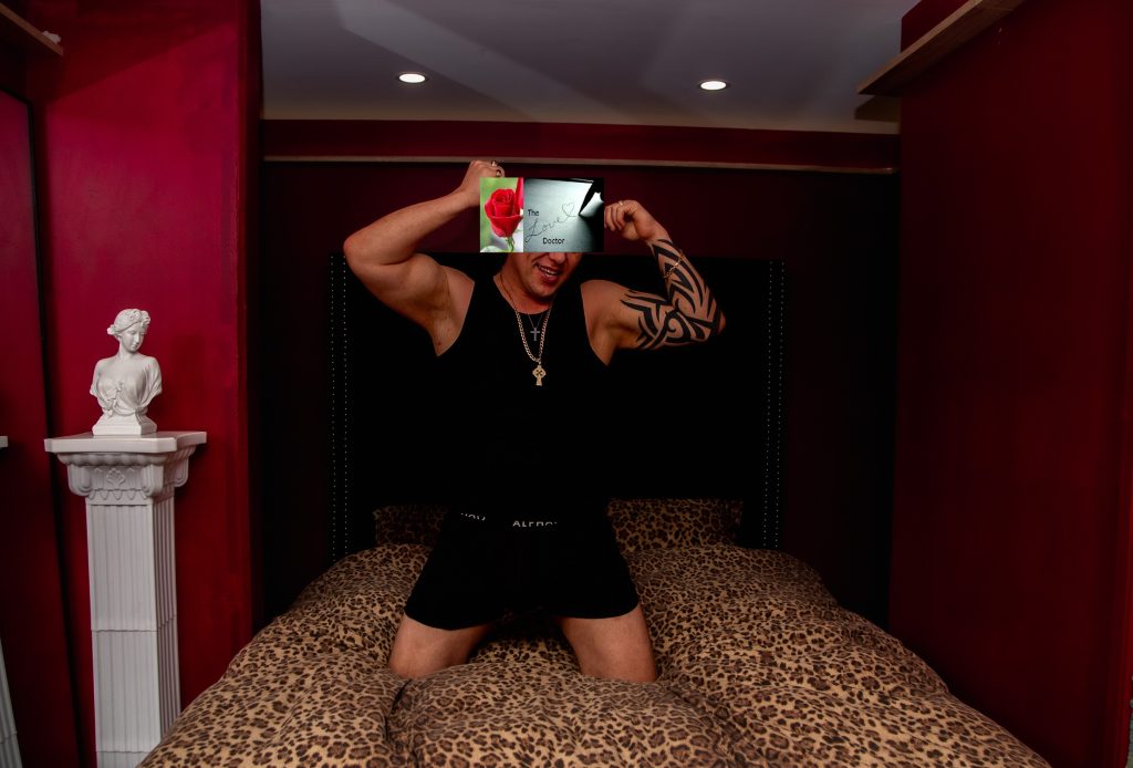 Man in black tank top and boxers flexes on a leopard print bed, holding a tablet with a rose image.