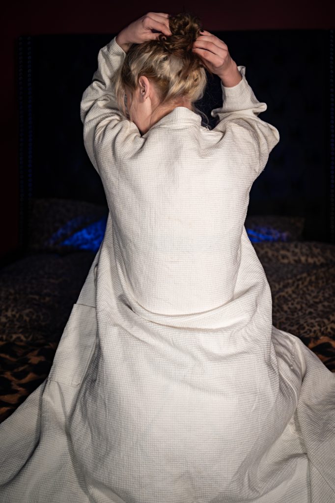 Woman in white robe tying hair on leopard print bed, July 2026 gallery.