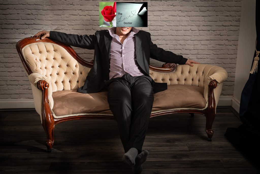 Man in suit on chaise lounge with 'The Love Doctor' book cover over face